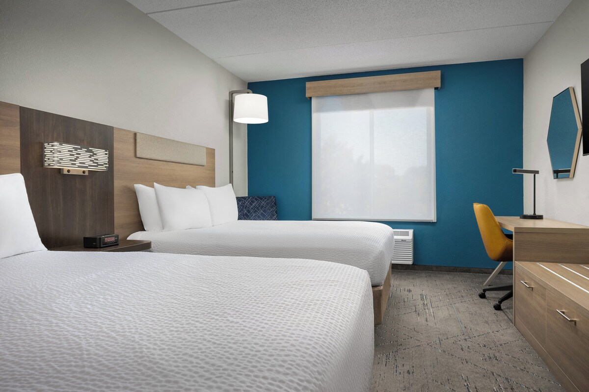 Quarto de hotel com duas camas, uma mesa, uma cadeira, um abajur, um aquecedor e uma janela grande.