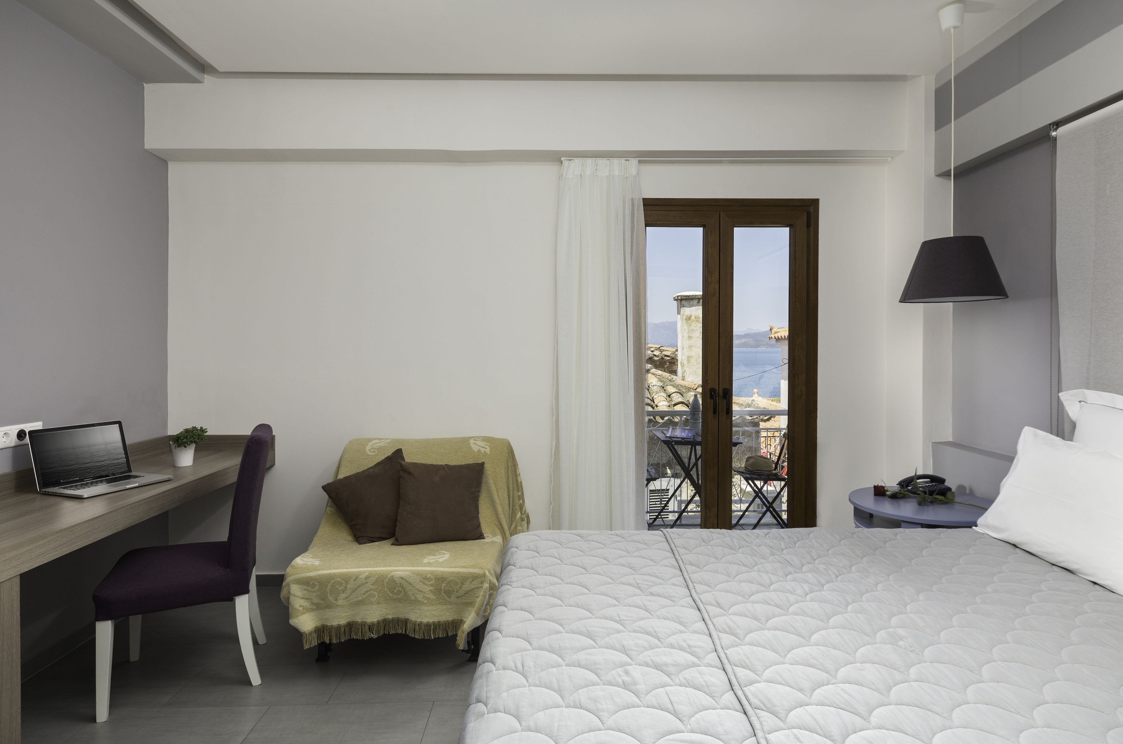A hotel room with a bed, a desk with a laptop, a chair, a small table, and a balcony with a view of the sea.