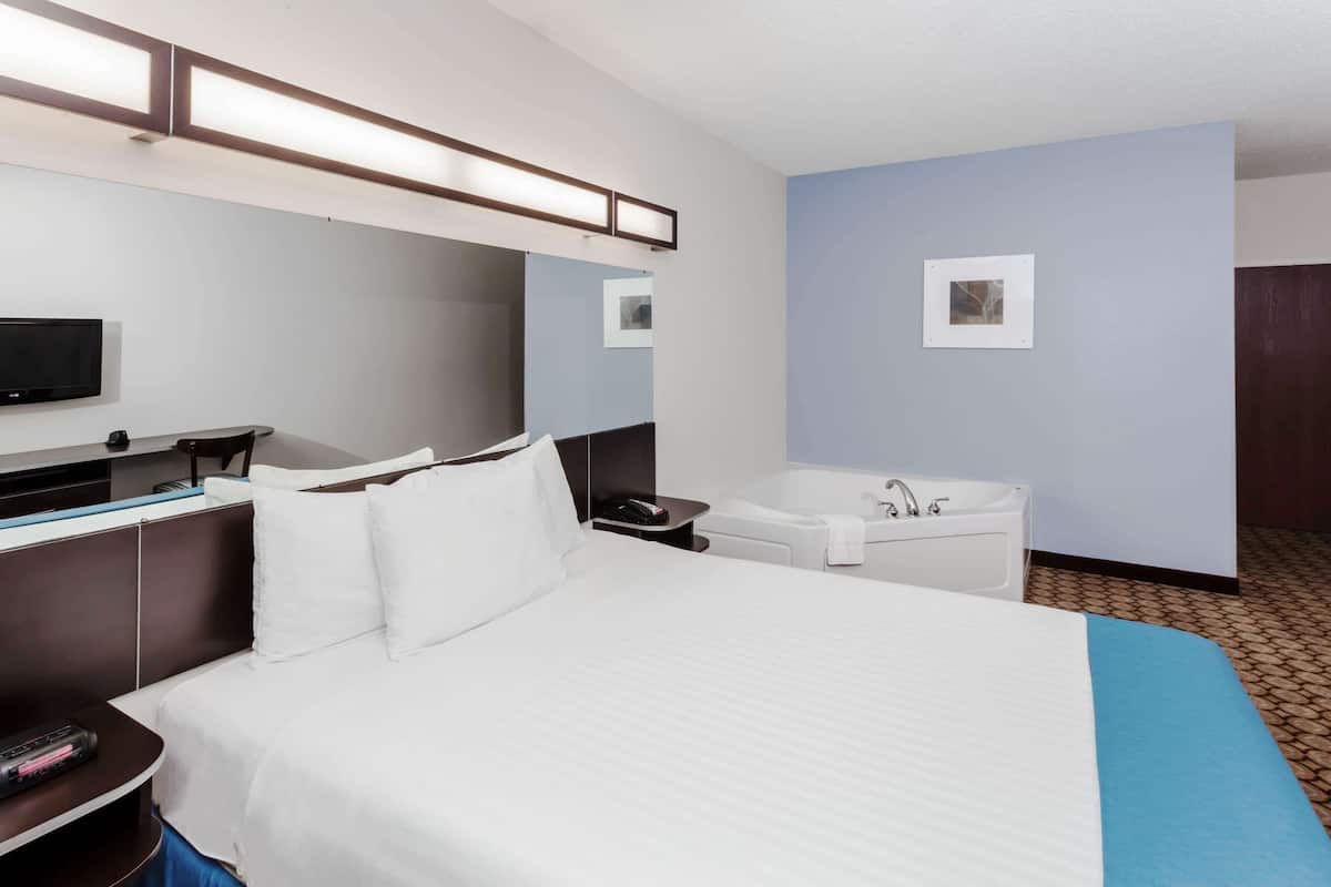 A hotel room with a large bed, a flat-screen TV, a desk with a chair, a desk lamp, a wall-mounted mirror, a bathtub, and a small framed picture on the wall.