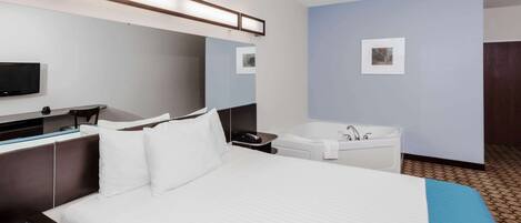 A hotel room with a large bed, a flat-screen TV, a desk with a chair, a desk lamp, a wall-mounted mirror, a bathtub, and a small framed picture on the wall.