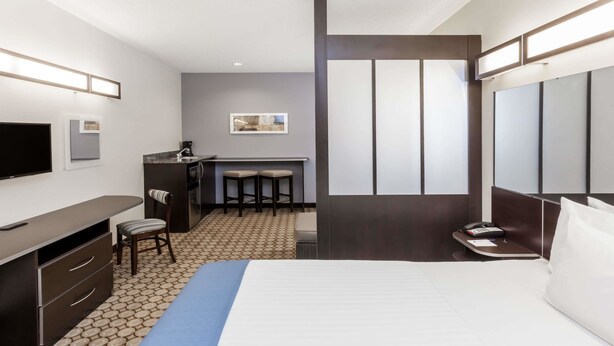 A hotel room with a bed, a desk, a chair, a TV, a minibar, and a small kitchen area with stools.