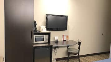 A compact hotel room with a kitchenette, a microwave, a coffee maker, a flat-screen TV, a small refrigerator, a chair, and a bed with a blue cover.