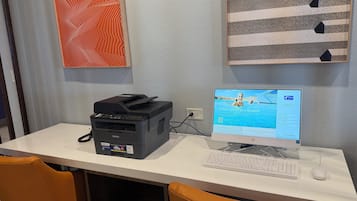 Un escritorio con una impresora Brother, una computadora de escritorio y un teclado. Hay dos sillas, una naranja y una marrón.