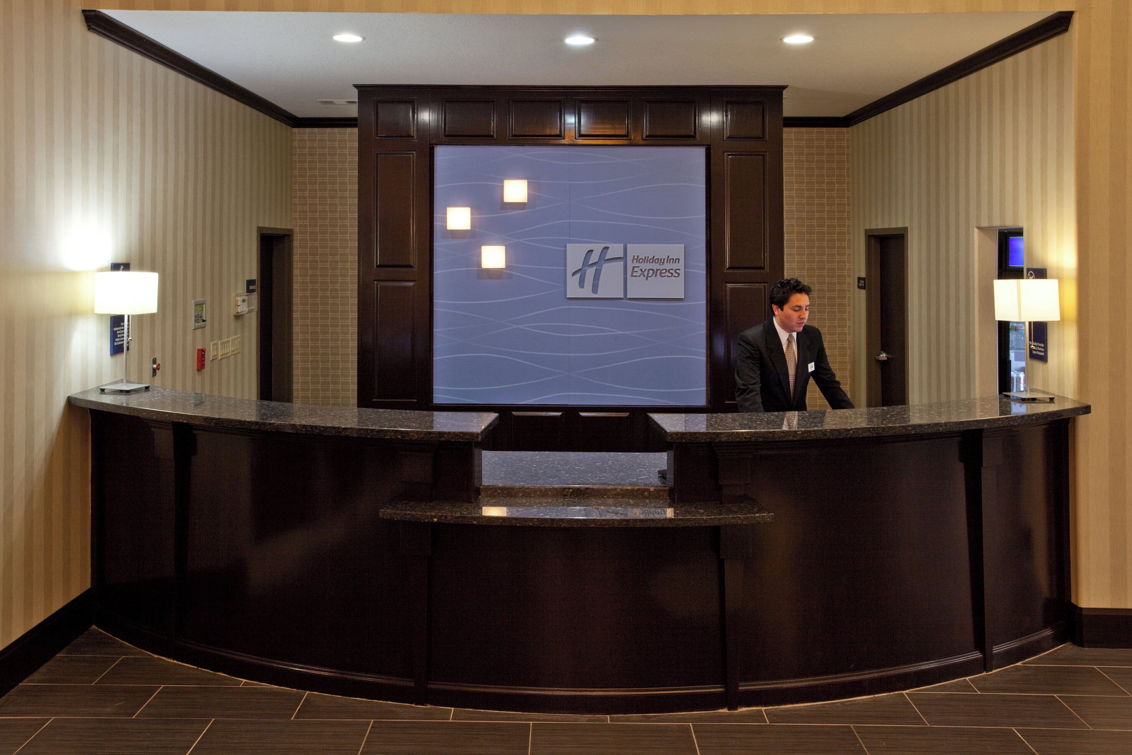 A reception desk with a digital screen displaying the Holiday Inn Express logo, a man standing behind it, and a wall-mounted light fixture to the left.
