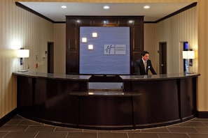 A reception desk with a digital screen displaying the Holiday Inn Express logo, a man standing behind it, and a wall-mounted light fixture to the left.