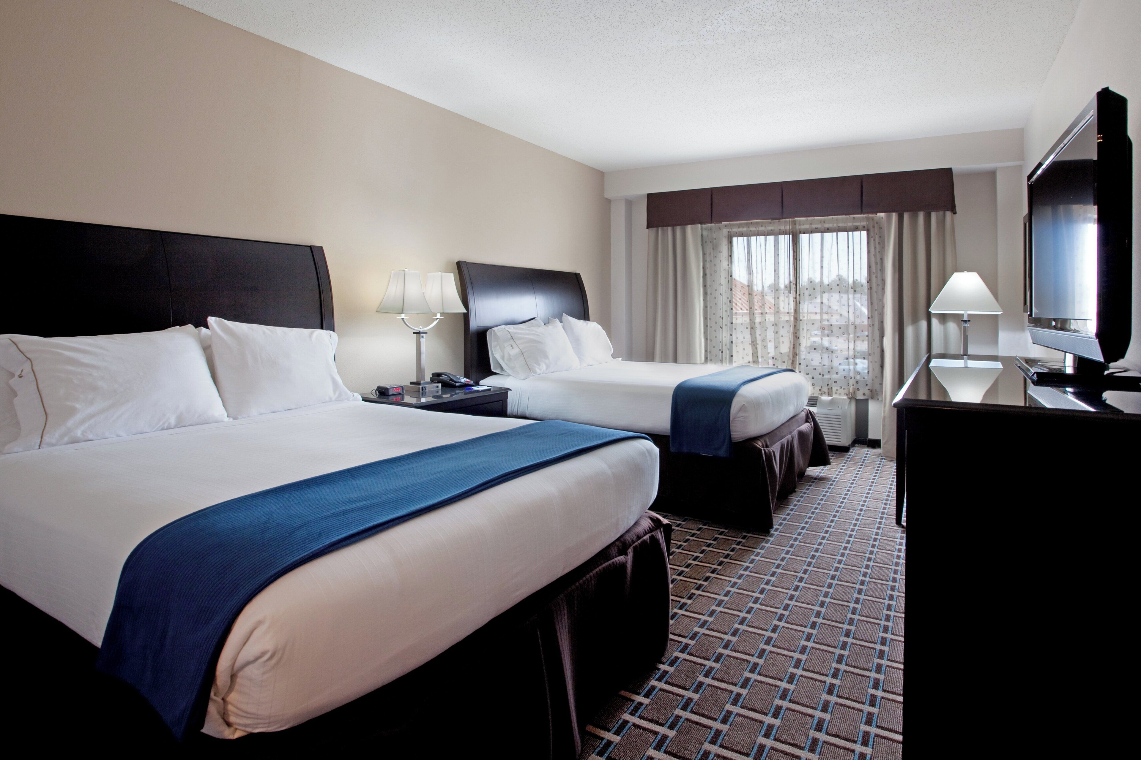 A hotel room with two beds, a desk, a TV, and a window with curtains.