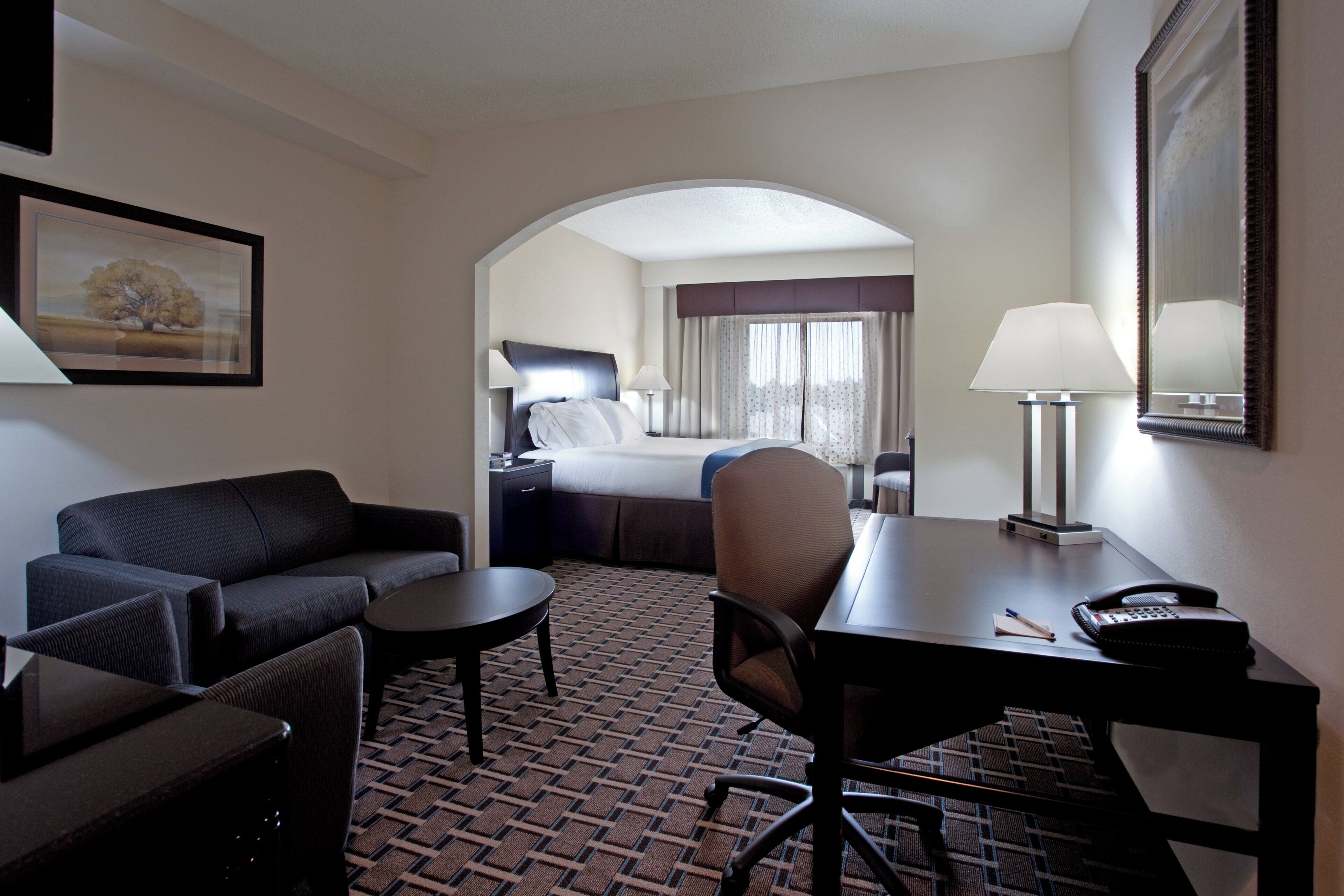 A hotel room with a sofa, a small table, a desk with a chair, a bed with a nightstand, and a lamp.