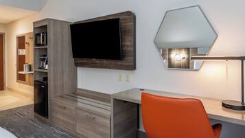 A hotel room with a flat-screen TV mounted on the wall, a desk with a chair, a lamp, and a built-in bookshelf.