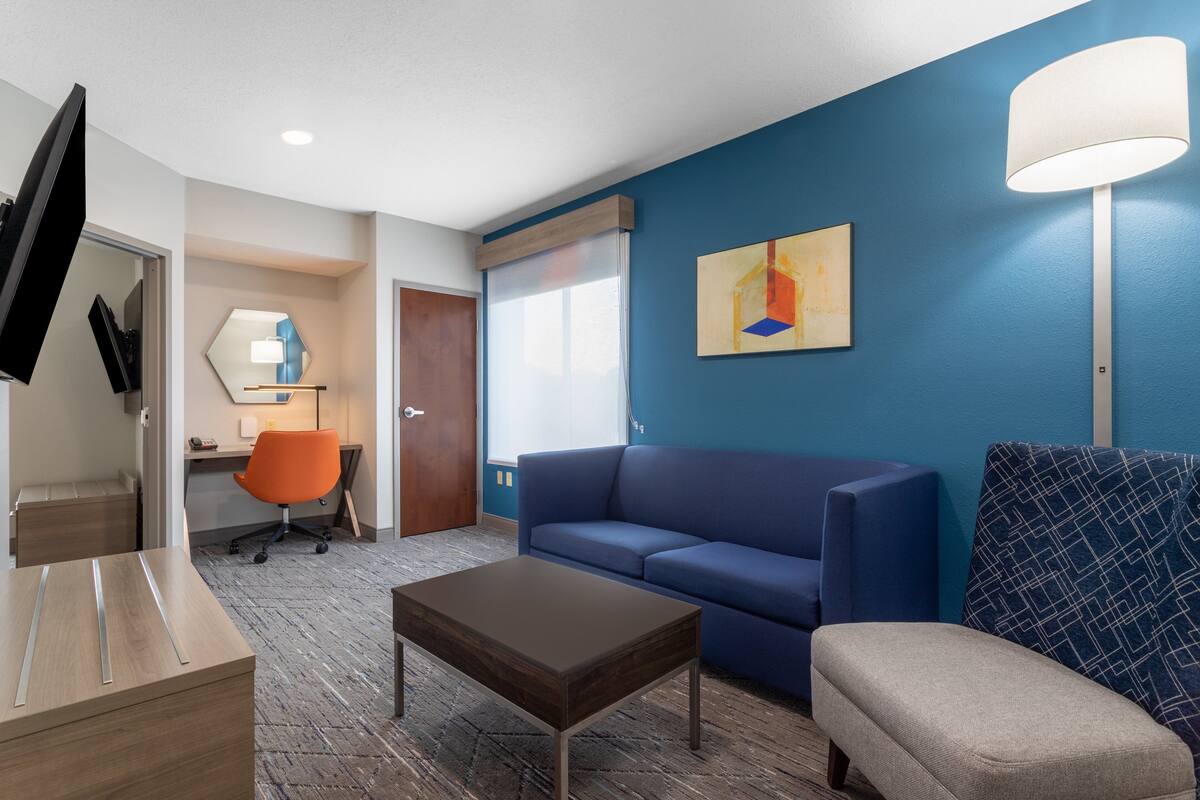 A modern hotel room with a blue accent wall, a sofa, a coffee table, a desk with a chair, and a TV.