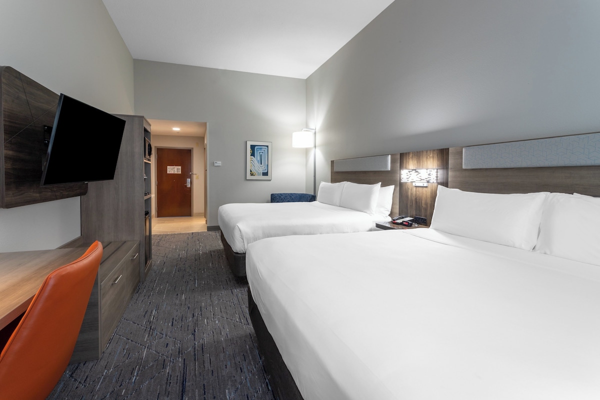 A hotel room with a desk, two beds, a TV, and a painting on the wall.