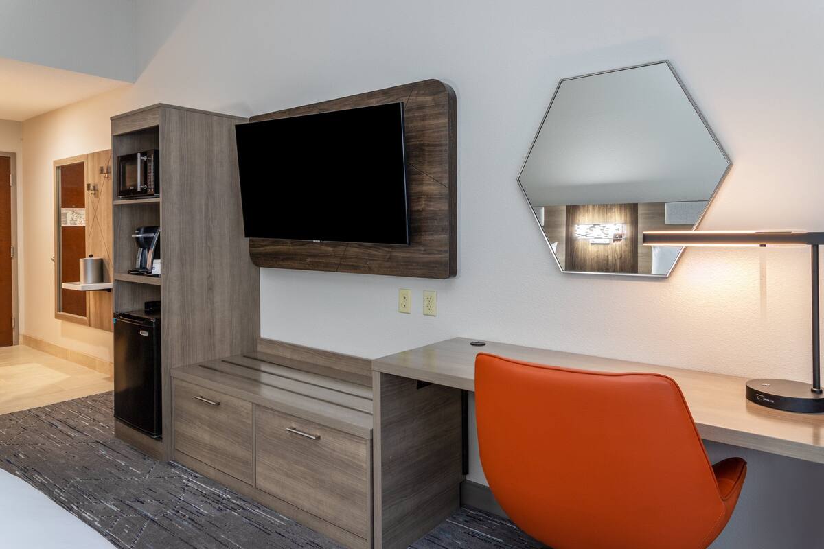 A modern hotel room with a wooden desk, a flat-screen TV, a built-in bookshelf, a coffee machine, and a bright orange chair.