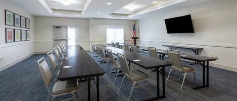 A conference room with long tables and chairs, a flat-screen TV mounted on the wall, and an American flag.