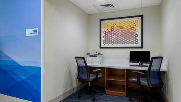 Un centre d’affaires doté d’un bureau blanc, de deux chaises de bureau noires, d’une œuvre d’art abstraite encadrée et d’un panneau indiquant que les locaux sont accessibles 24 heures sur 24.