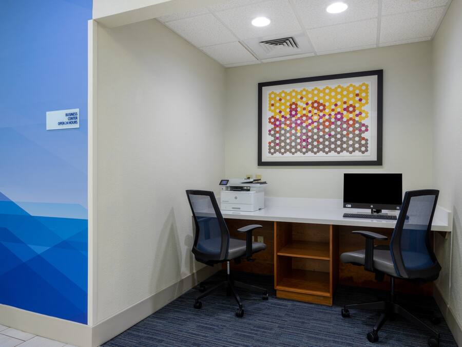 A business center with a white desk, two black office chairs, a framed abstract art piece, and a sign indicating 24-hour availability.