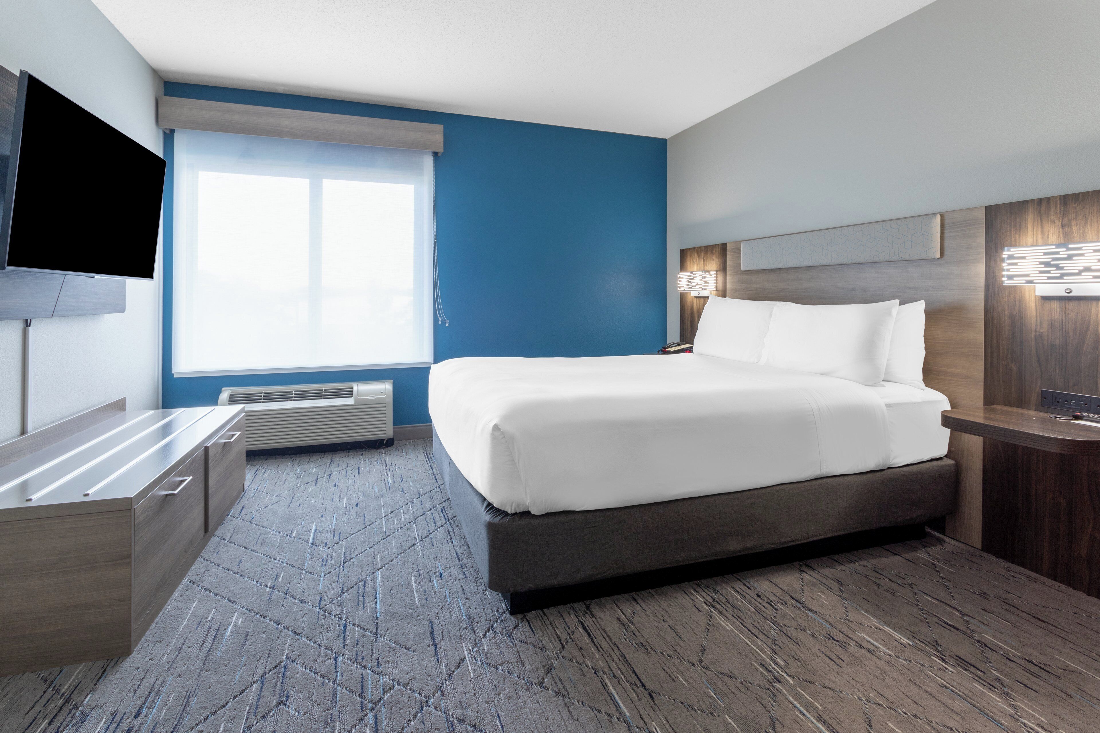 Ein Hotelzimmer mit einem großen Bett, einem Flachbildfernseher, einem Schreibtisch und einem Fenster mit Jalousien.