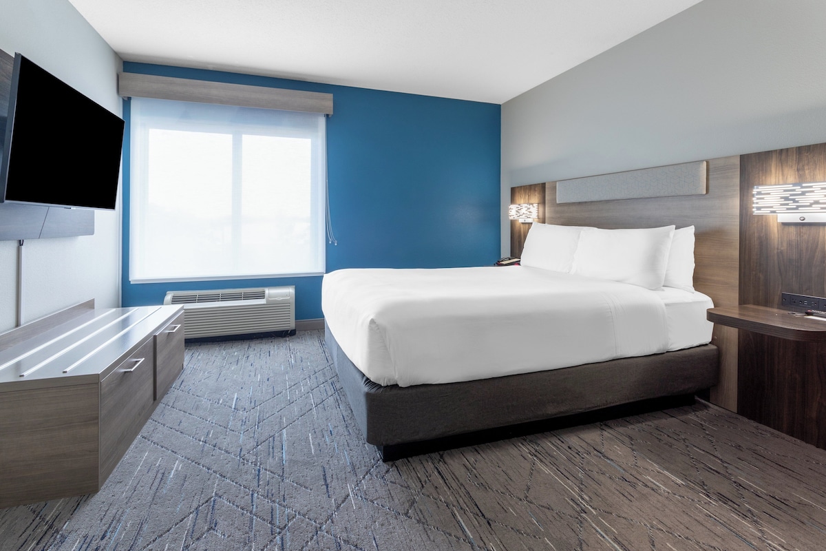 A modern hotel room with a large bed, a flat-screen TV, a desk, and a window with blinds.