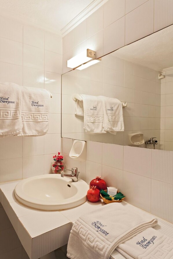 A hotel bathroom with a large mirror, white tiled walls, a sink, and multiple towels with "Hotel Donyssos" branding. - Hotel Dionyssos (Skopelos)