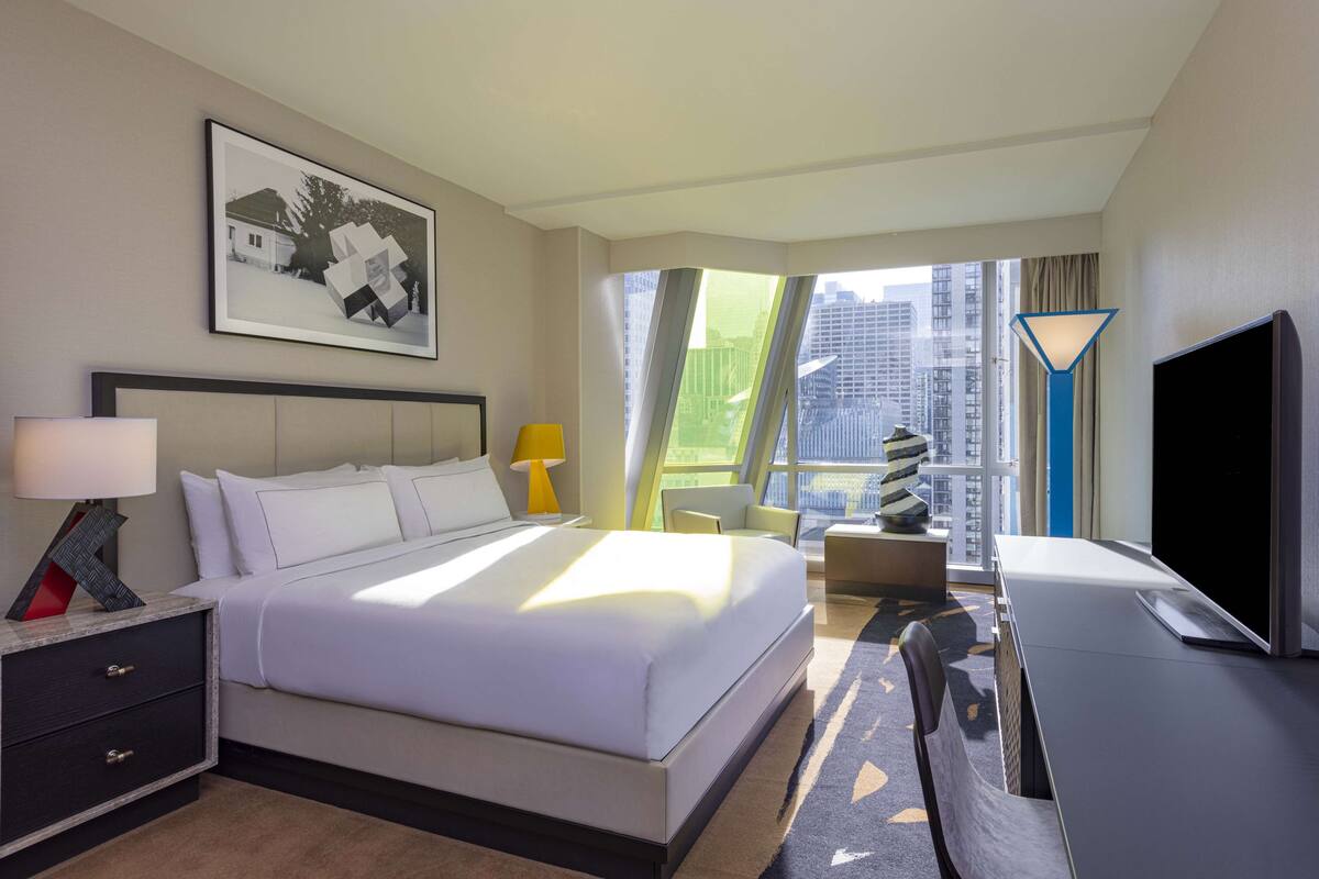 A hotel room with a large bed, a desk with a flat-screen TV, a chair, a nightstand, a lamp, and a window with a city view.