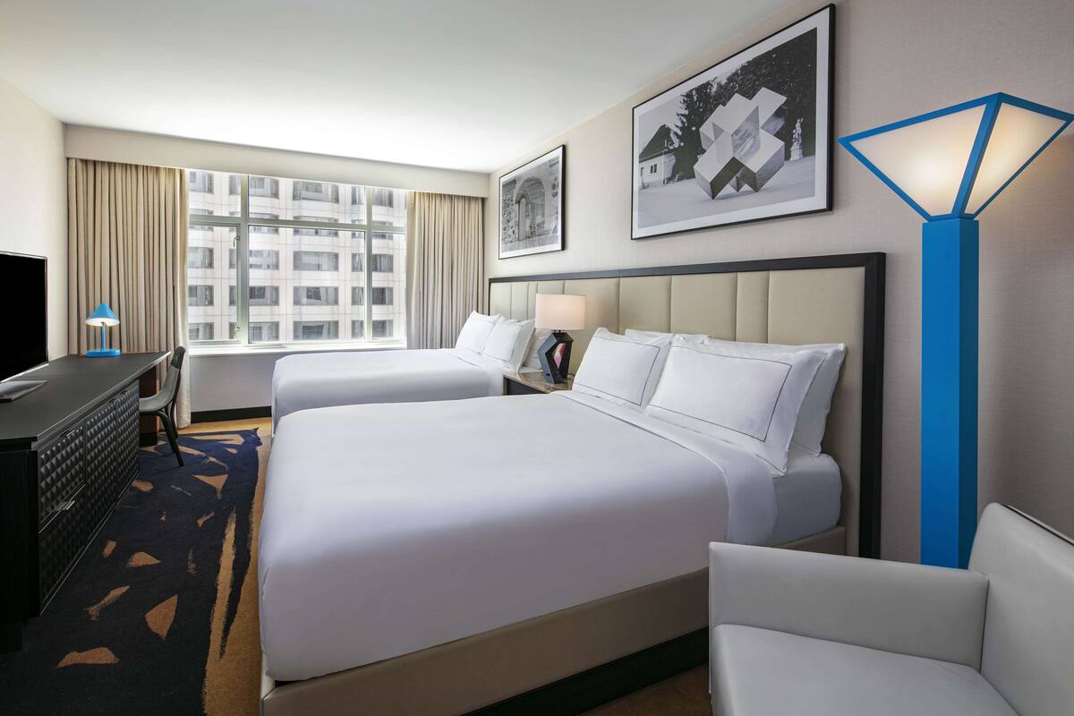 A hotel room with two beds, a desk, a chair, a lamp, and a large window with a view of buildings.