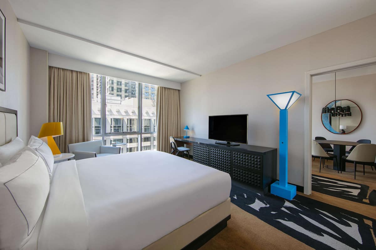 A hotel room with a large bed, a desk with a chair, a television, a blue floor lamp, and a dining area with chairs and a table.