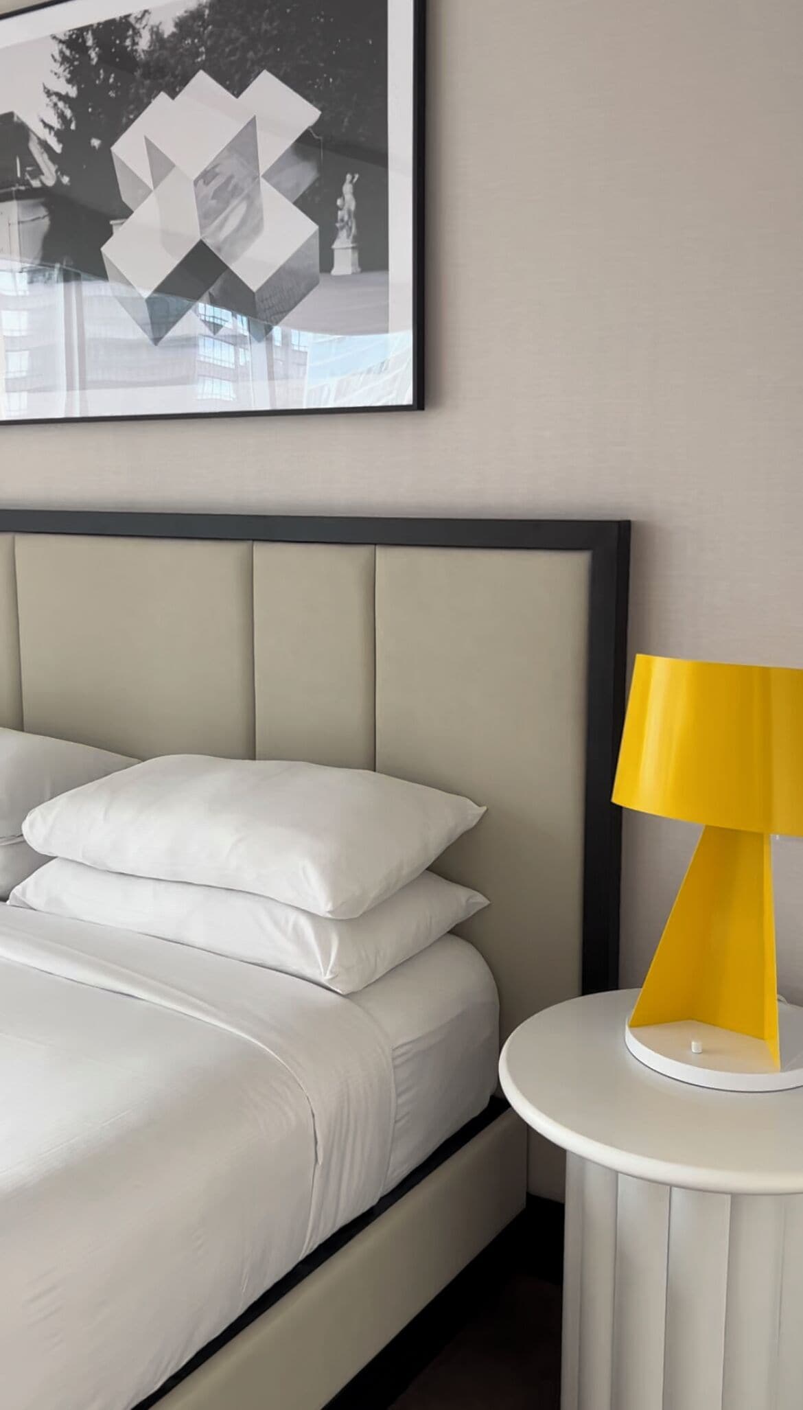 A neatly made bed with white linens and pillows, a modern yellow lamp on a small white table beside it, and a geometric abstract painting on the wall.