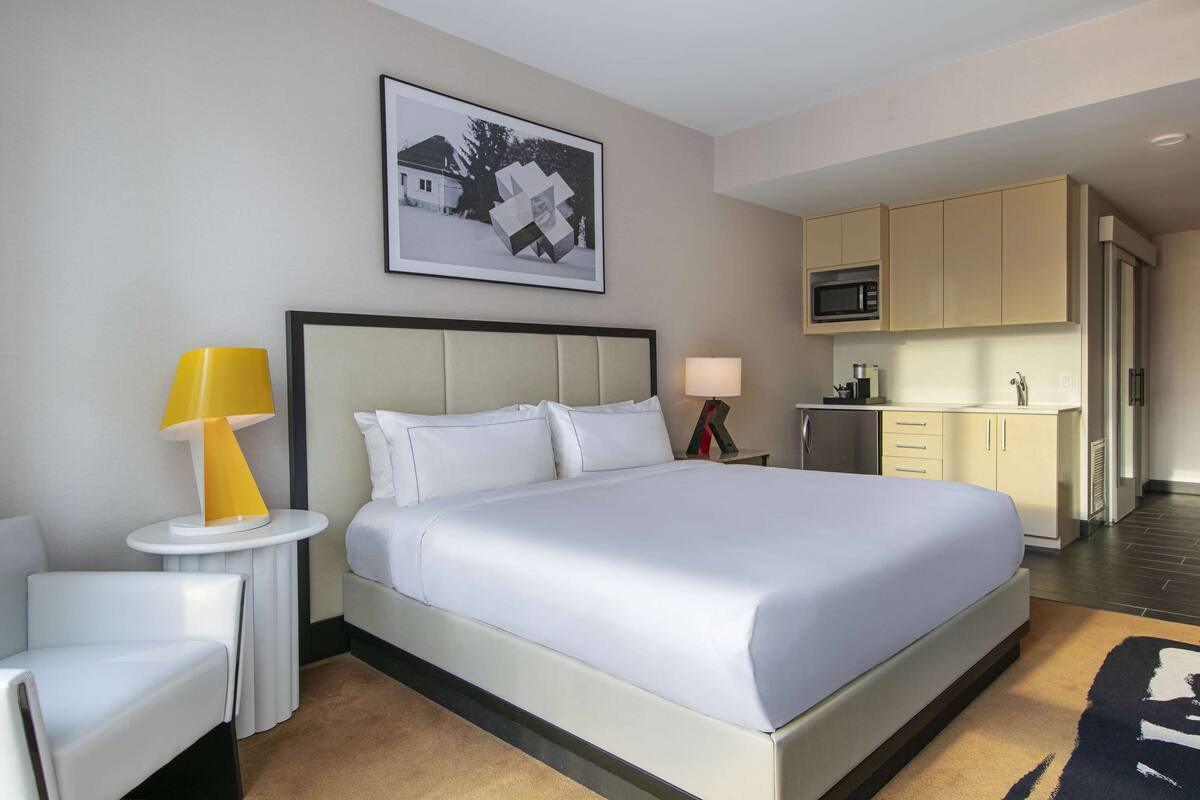 A hotel room with a large bed, a bedside table with a lamp, a chair, a kitchen area with a microwave, and a framed picture on the wall.