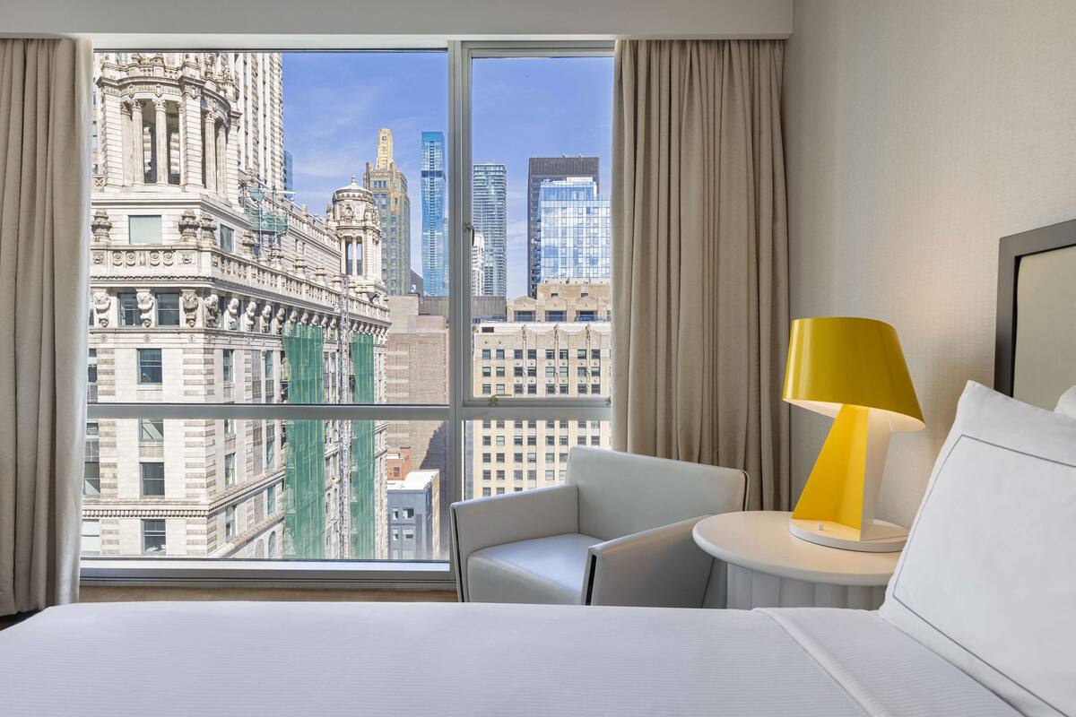 A hotel room with a large window offering a city view, a white bed, a white armchair, and a small table with a yellow lamp.