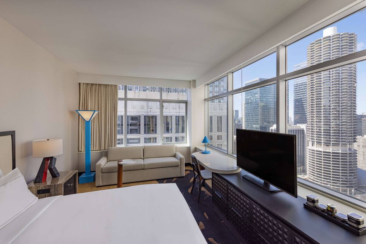 A hotel room with a large bed, a TV, a sofa, and a desk. There is a large window with a view of tall buildings.