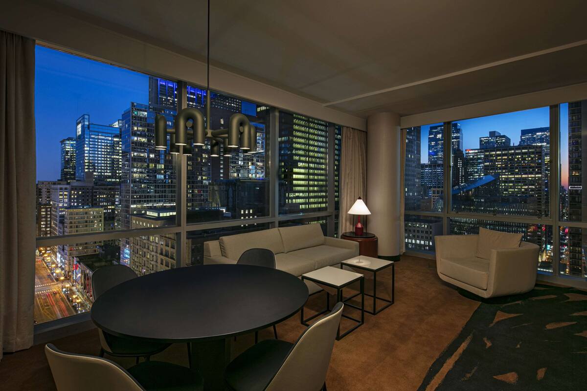 A modern hotel room with a large city view, a round dining table, two sofas, and a lamp.
