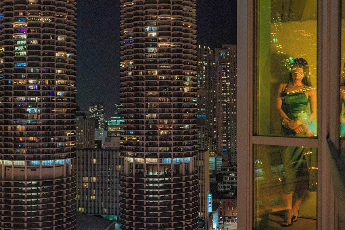 A nighttime cityscape with tall buildings and a reflection of a person in a window.