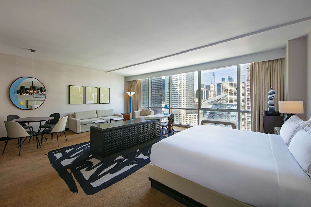 A hotel room with a large bed, a desk, a chair, a sofa, a coffee table, and a view of the cityscape.