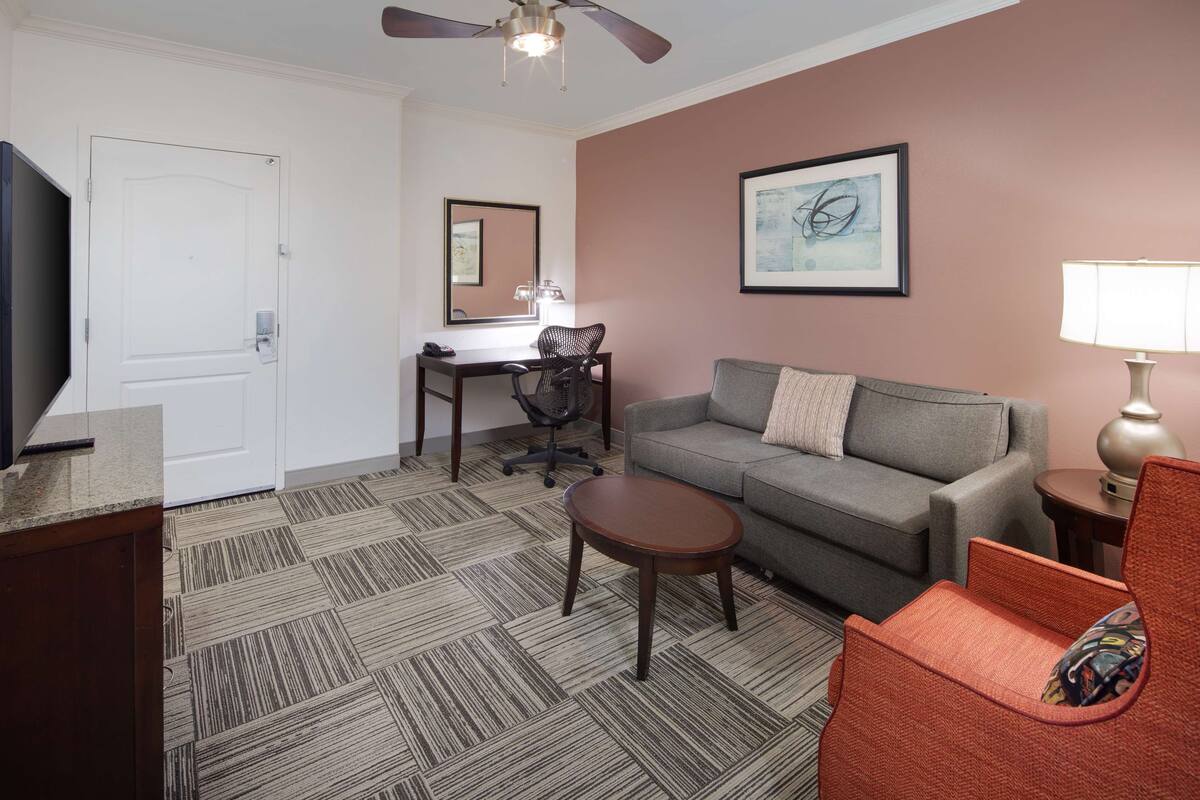 A hotel room with a gray sofa, a small round table, a desk with a chair, a TV, and a lamp.
