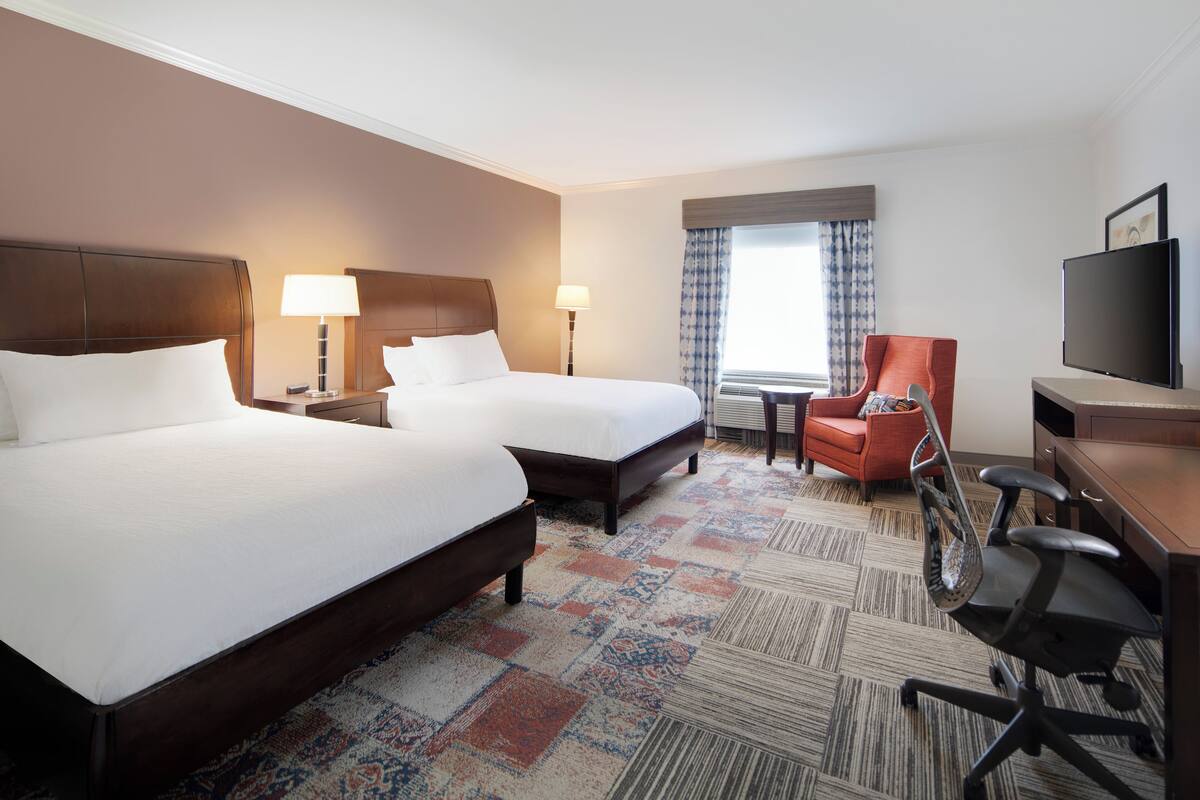 A hotel room with two beds, a desk, a chair, a TV, and a window with curtains.