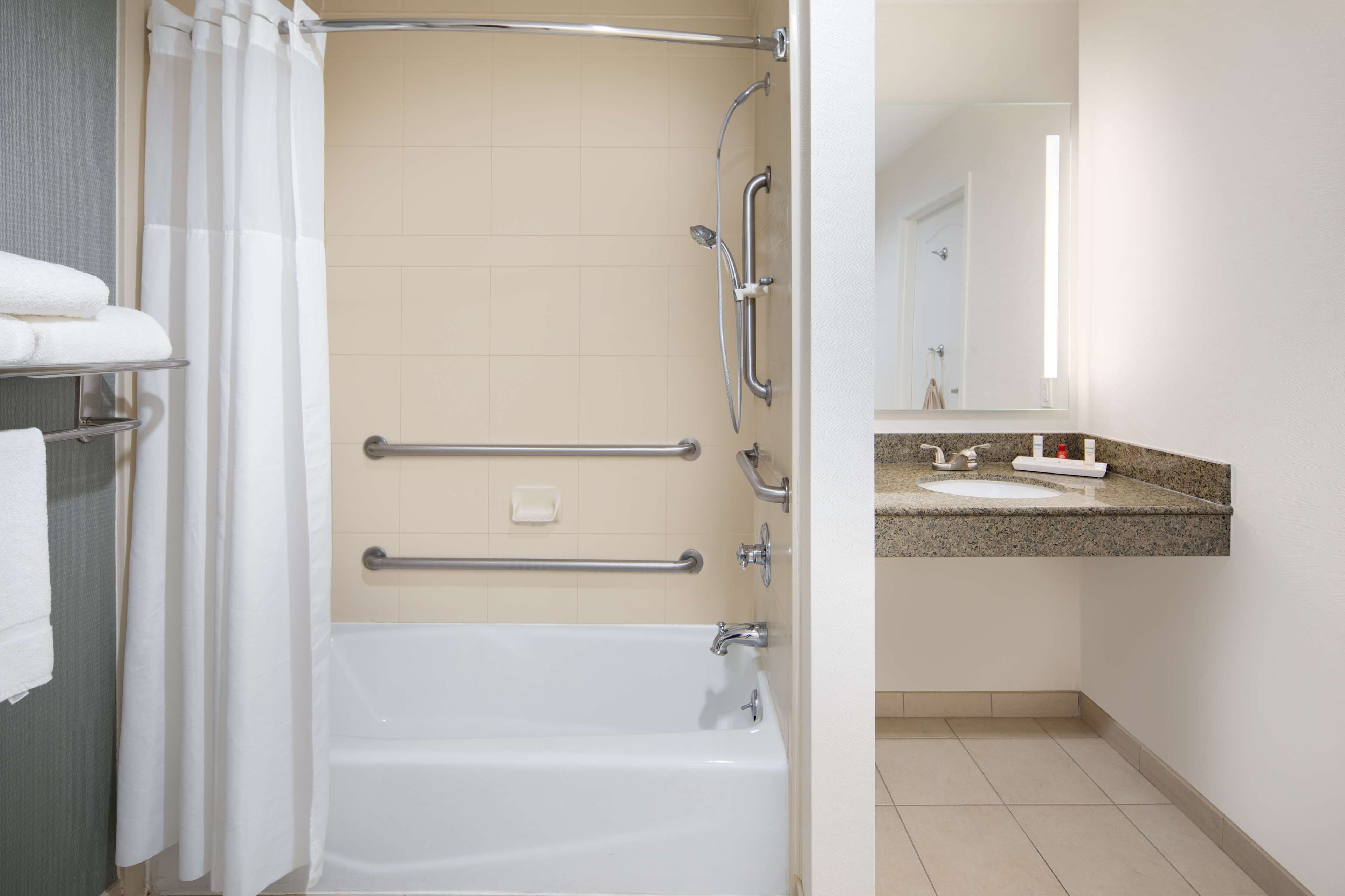 Un baño con bañera, barras de apoyo, ducha, lavabo y espejo.