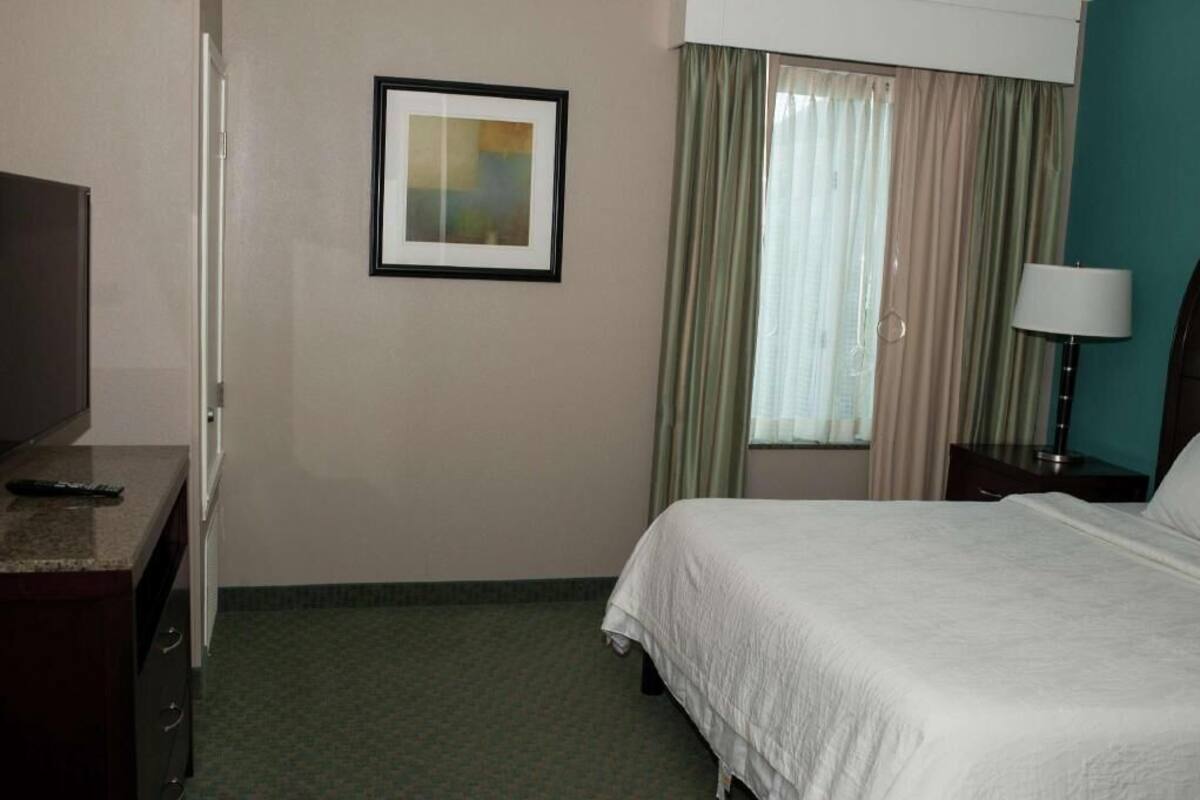 A hotel room with a bed, a desk with a remote control, a framed picture on the wall, a window with curtains, and a lamp on a nightstand.
