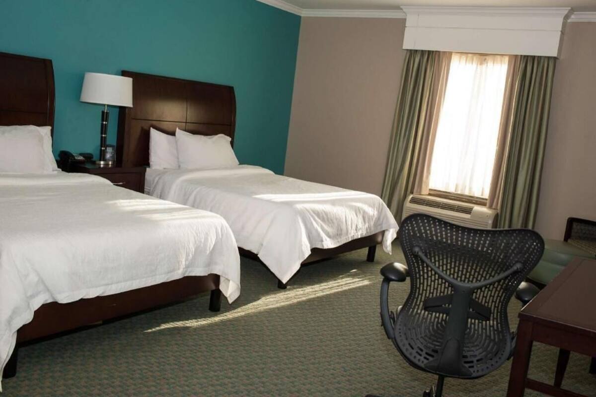 A hotel room with two beds, a desk, a chair, a lamp, and a window with curtains.