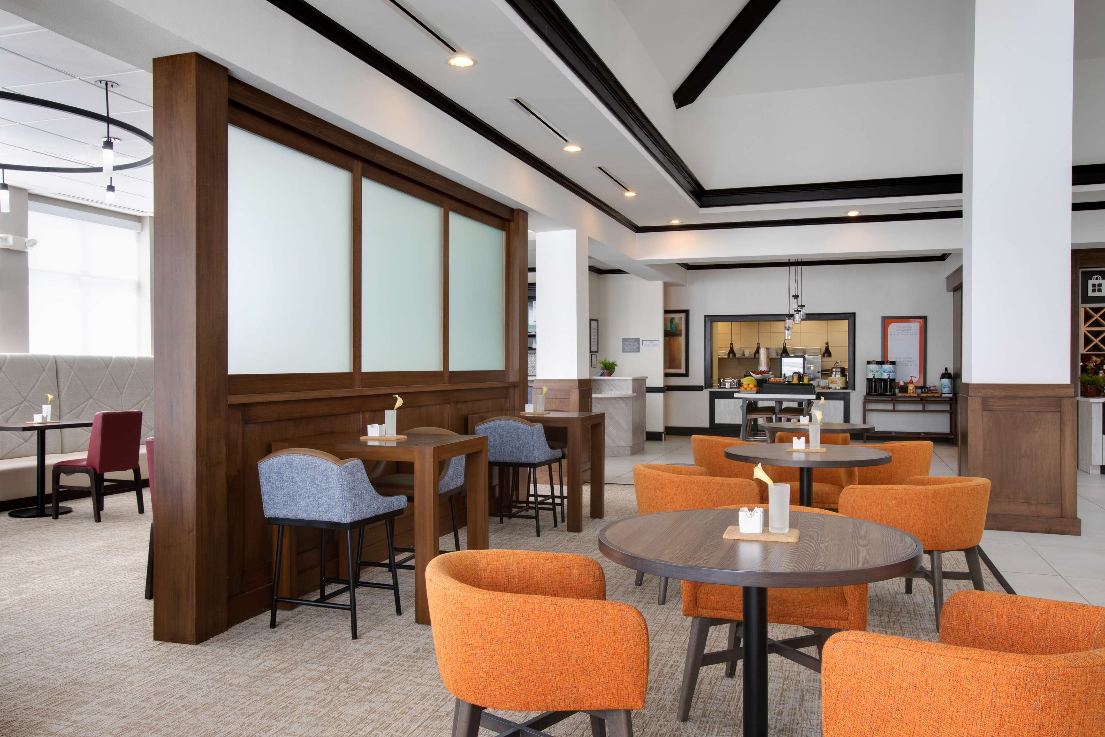 Un lobby moderno de hotel con mesas redondas y sillas anaranjadas, una zona de bar y vistas a la cocina.