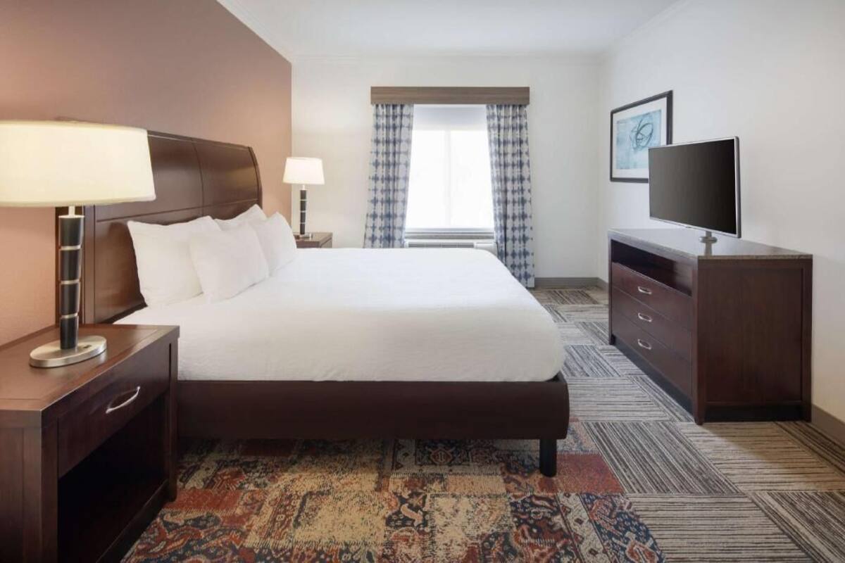 A hotel room with a large bed, a TV, a desk with a lamp, and a patterned carpet.