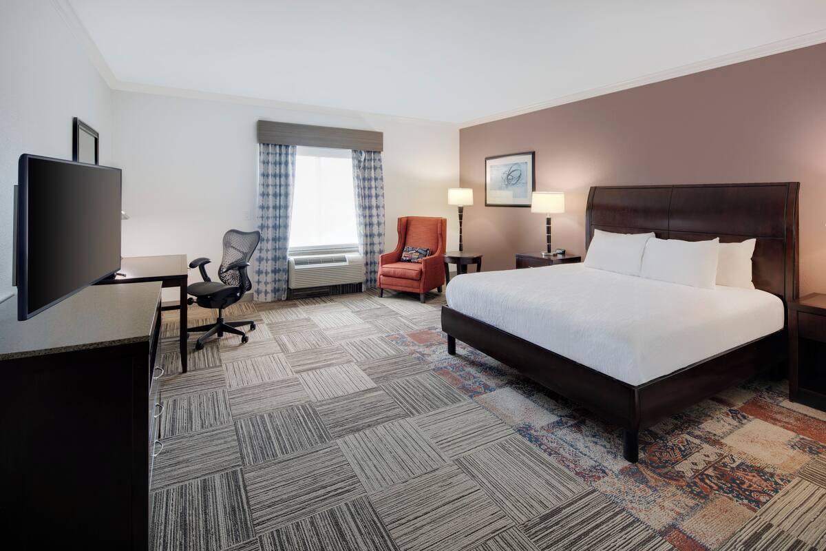 A hotel room with a large bed, a desk with a chair, a television, and a window with curtains.