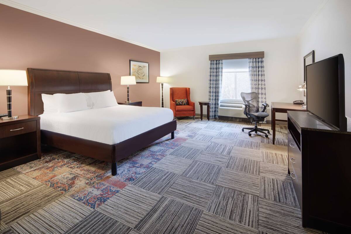 A hotel room with a large bed, two bedside tables with lamps, a desk with a chair, a television, and a window with curtains.