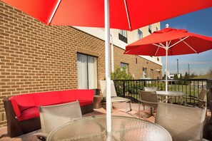 An outdoor seating area with a red sofa, wicker chairs, and a glass table, under red umbrellas, with a brick building in the background. - Holiday Inn Express Hotel & Suites Columbia East - Elkridge by IHG (Elkridge)