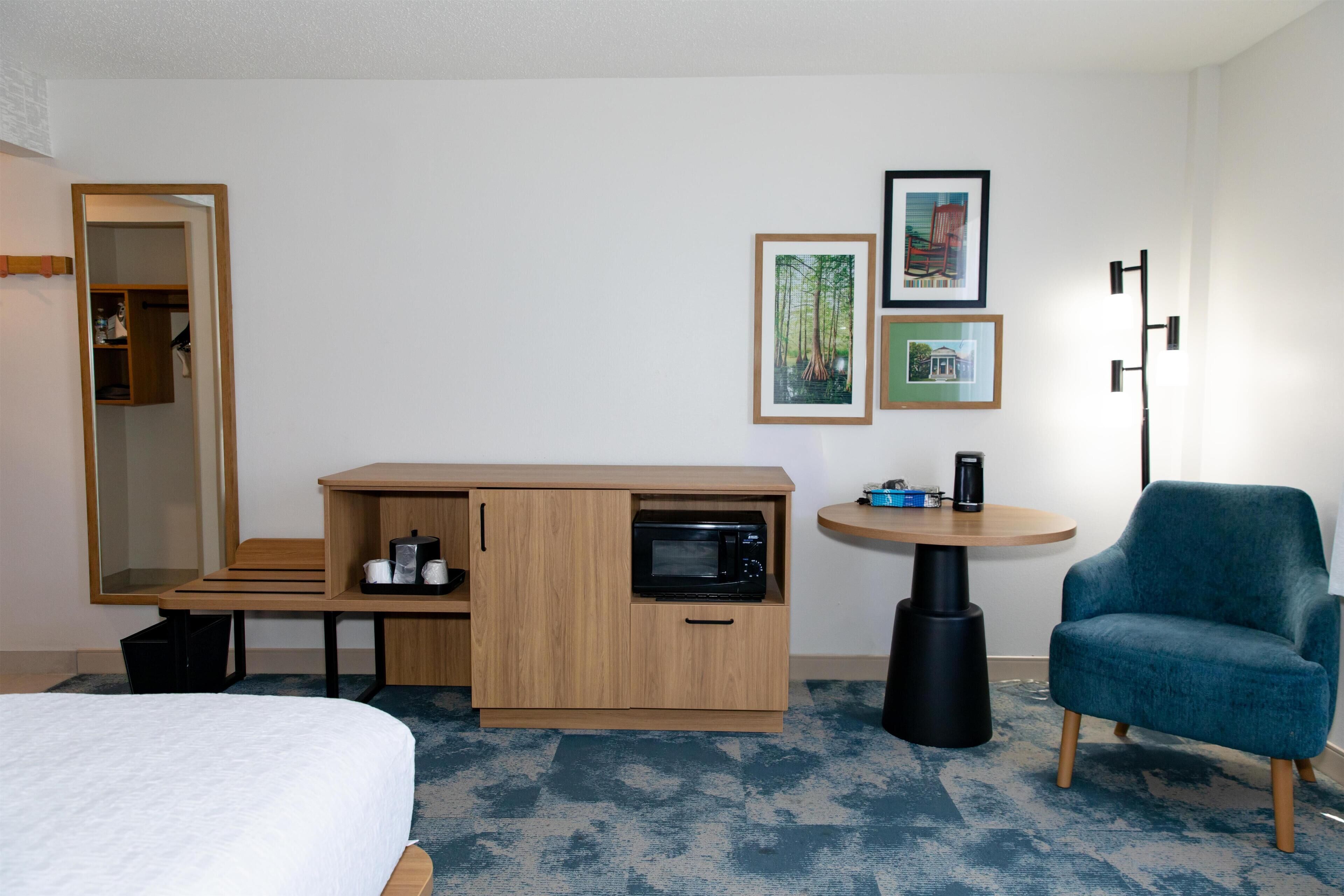 Una camera d'albergo con un letto, una scrivania con un forno a microonde, una sedia e un tavolino con una lampada. Alle pareti sono presenti quadri incorniciati.