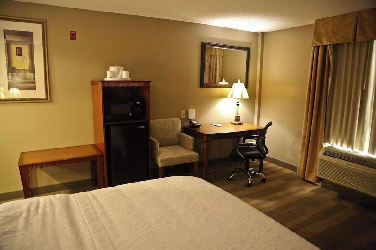 A hotel room with a bed, a desk, a chair, a small cabinet, a lamp, a mirror, and a window with curtains.