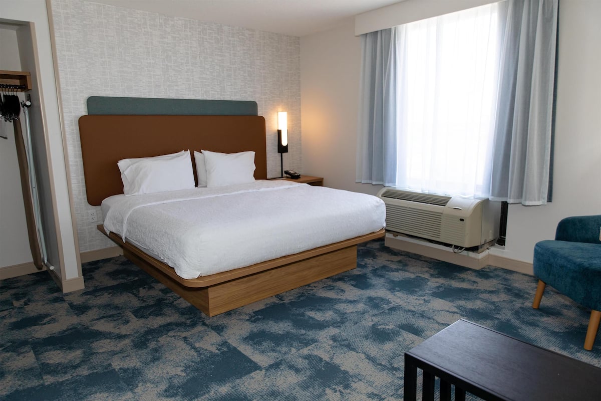 A hotel room with a large bed, a bedside table, a chair, a window with curtains, and an air conditioning unit.