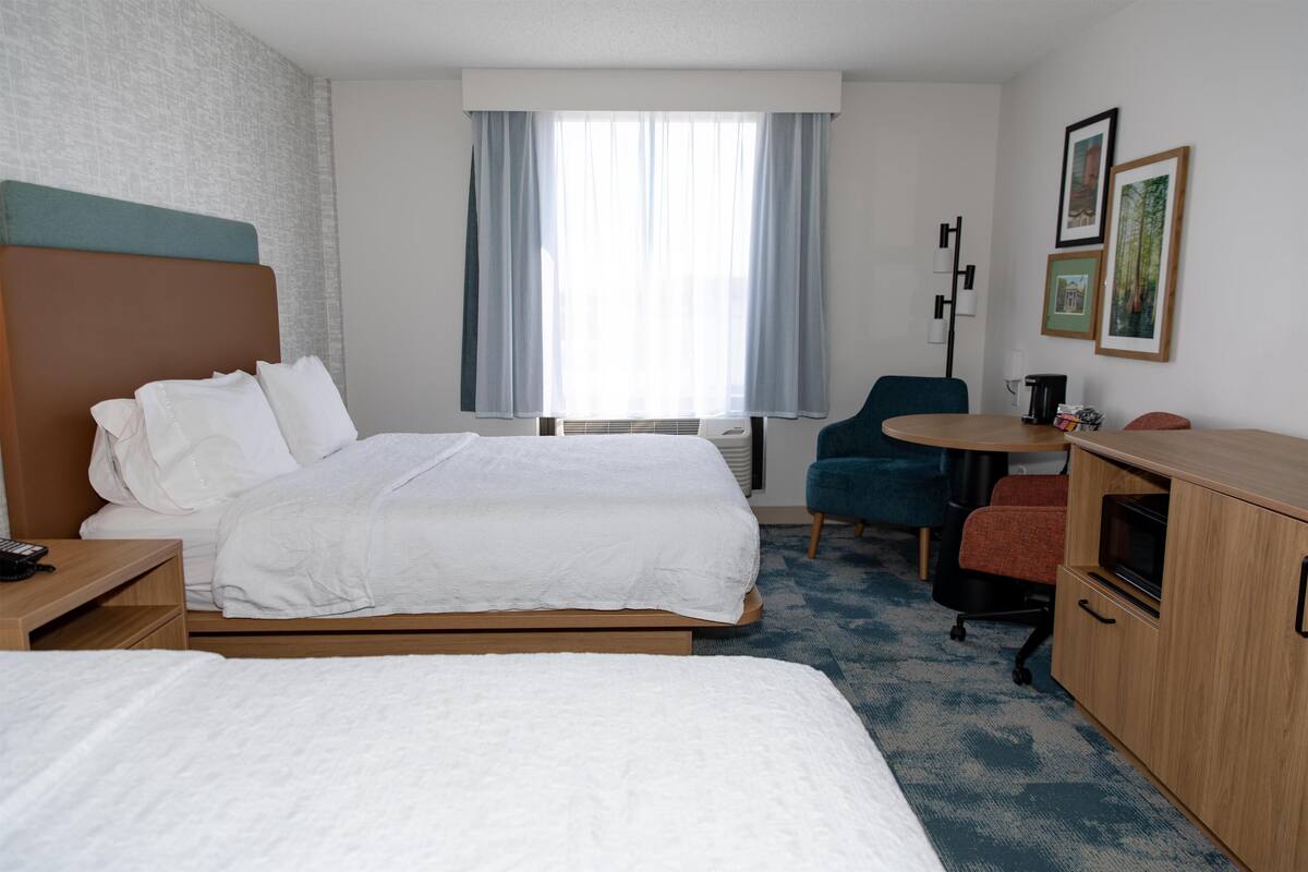 A hotel room with two beds, a desk, a chair, a small table, and a window with curtains.