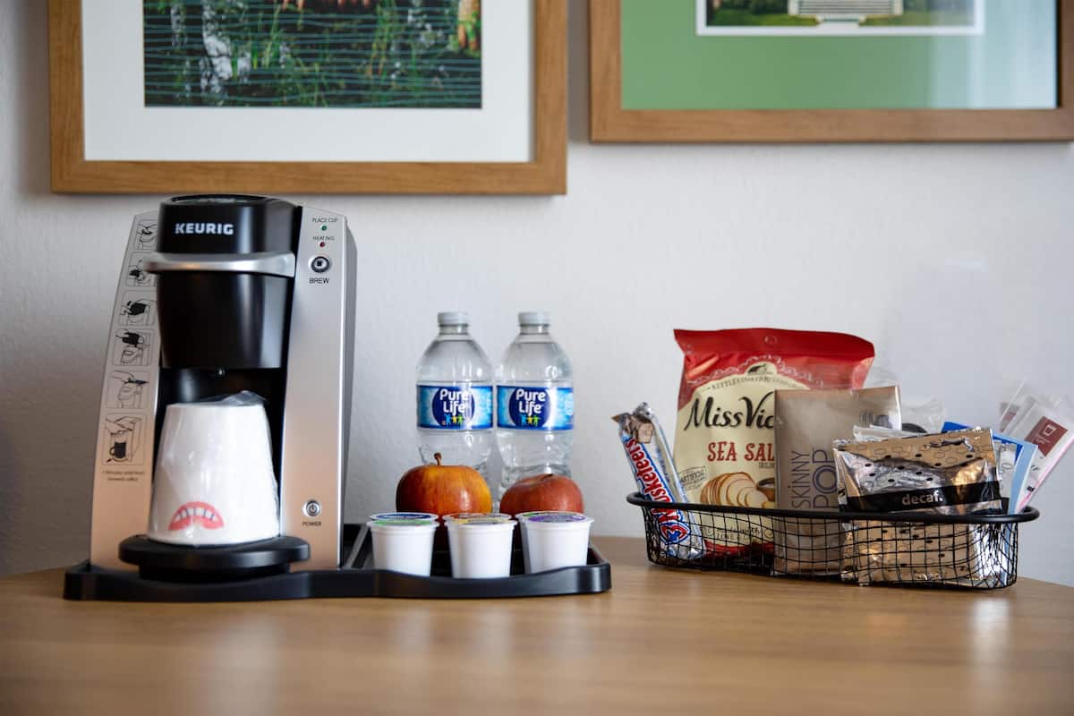 A Keurig coffee maker with a cup holder, two water bottles, two apples, a basket with snacks, and a bag of Miss Vicki's Sea Salt.