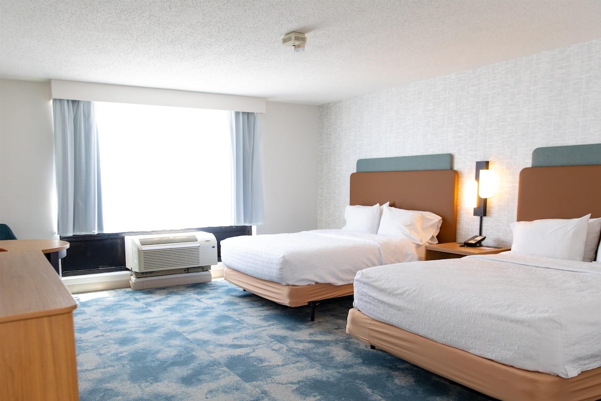 A hotel room with two beds, a desk, a chair, a window with curtains, an air conditioning unit, and a wall-mounted lamp.