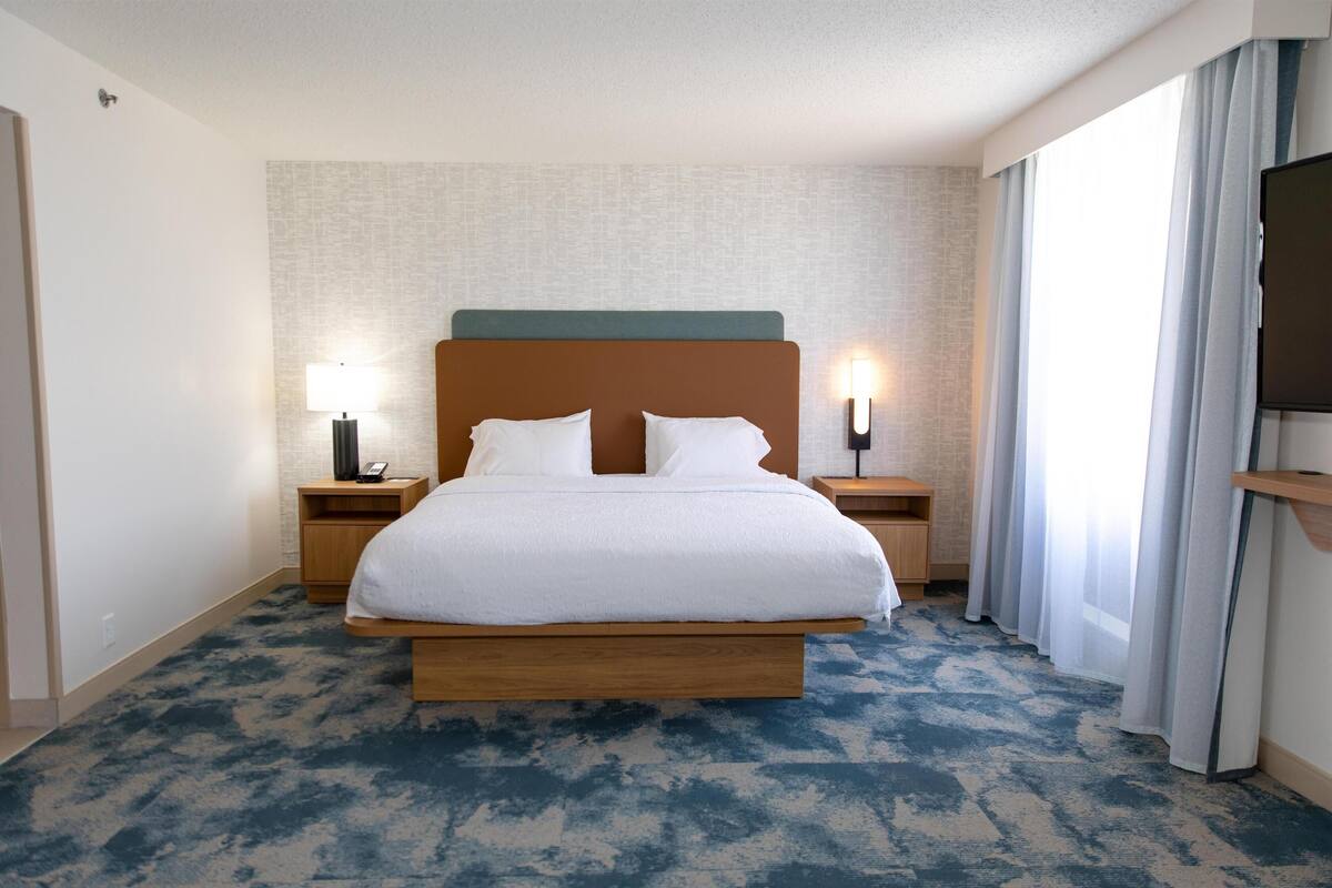 A hotel room with a large bed, two bedside tables with lamps, a flat-screen TV, and a large window with curtains.