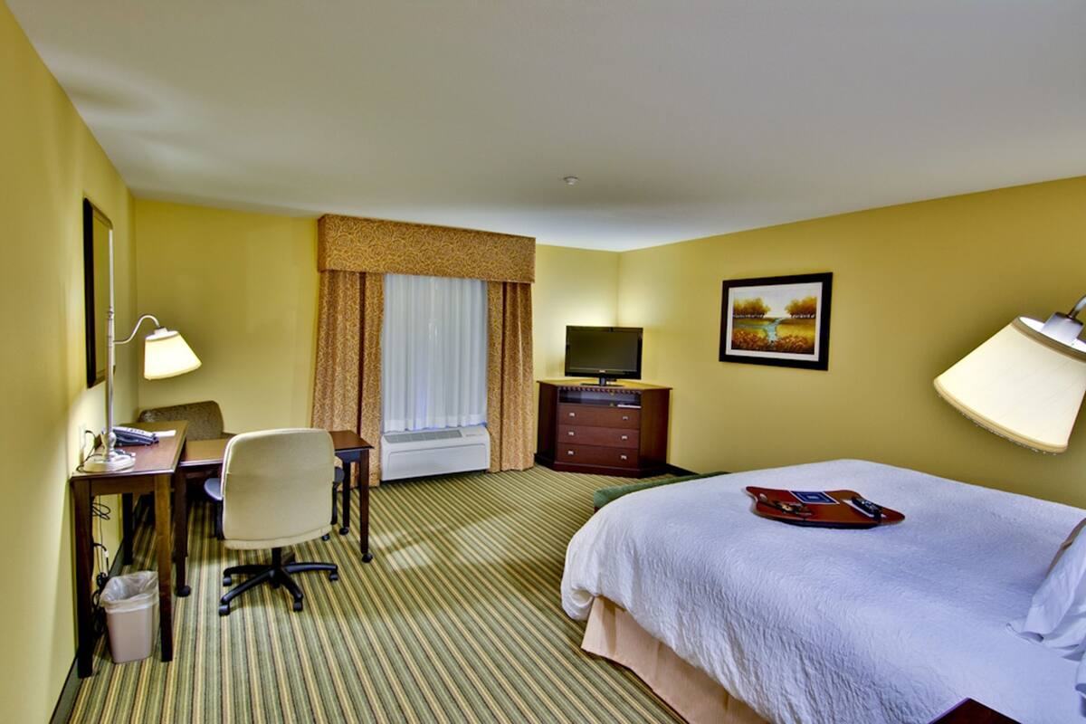 A hotel room with a bed, desk, chair, television, and a painting on the wall.