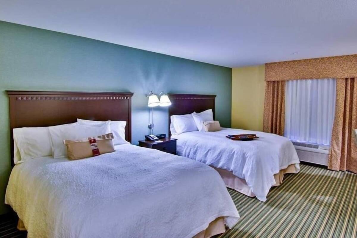 A hotel room with two beds, a nightstand with a lamp, and a window with curtains.